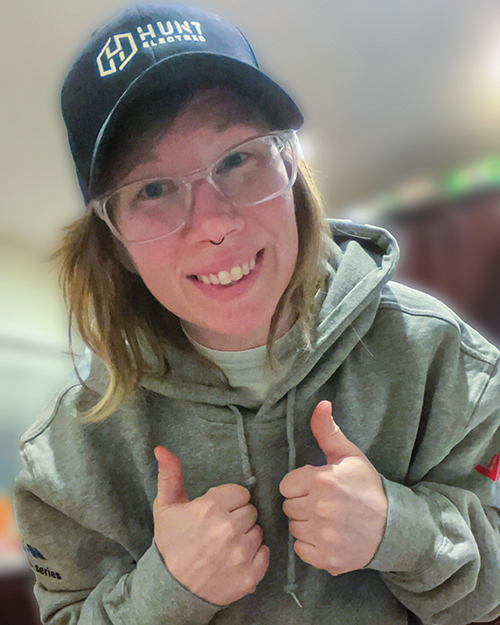 Electric apprentice Mandy Tullis-Martin wearing glasses, a 'Hunt Electric' hat, and a hoodie smiles while giving two thumbs up.