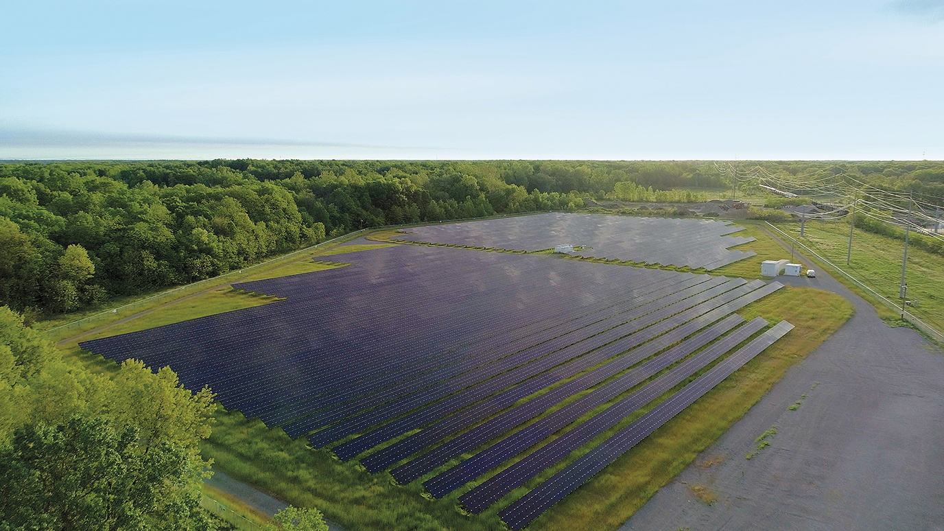 An aerial view of a large solar panel farm surrounded by trees, with power lines running along the right side of the field under a clear sky.