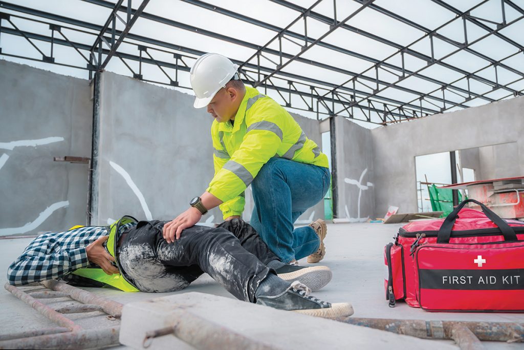 A construction worker in a yellow high-visibility jacket and hard hat is providing first aid to a coworker who is lying on the floor, injured. The worker kneels beside the injured person, checking on them. A red first aid kit is placed on the floor nearby. The scene takes place in a partially constructed building with exposed metal beams and unfinished walls.